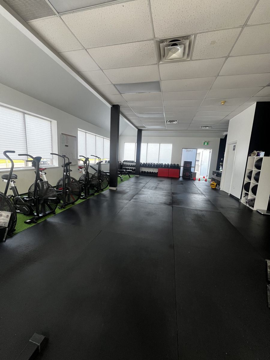 The open rubber training floor at TCT Gym in Oshawa, looking toward the Unit 108 entrance past air bikes and a dumbbell rack.